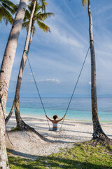 Fototapeta premium Woman on swing between palm trees on beach with ocean view