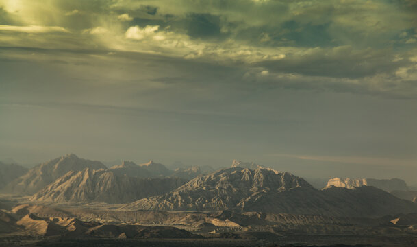 View of rugged, sun-baked mountains rise under a turbulent sky, casting shadows on the arid landscape, creating a stark yet captivating scene, Khuzdar, Pakistan.