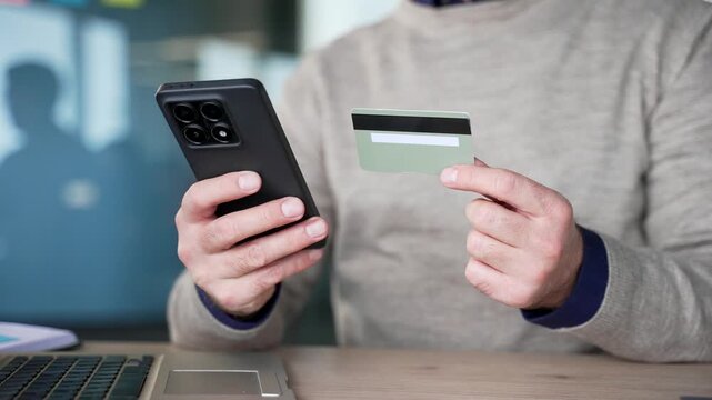 Close up of businessman hands holding credit card and using smartphone sitting at desk in office. Making online payment or shopping, using mobile phone for digital transaction and browsing services.