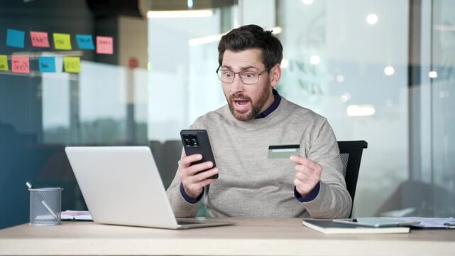 Shocked upset businessman checking credit card details on smartphone sitting at desk in modern office. Frustrated mature professional reacting to online fraud, facing scam or unauthorized transaction