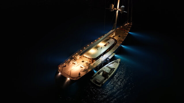 Aerial drone night shot of illuminated luxury yacht with wooden deck anchored in open ocean deep blue sea