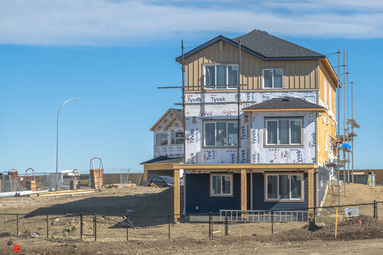 Calgary, Alberta, Canada. March 24, 2026. A modern two-story residential house under construction featuring Tyvek weather barrier and scaffolding on a dirt lot.