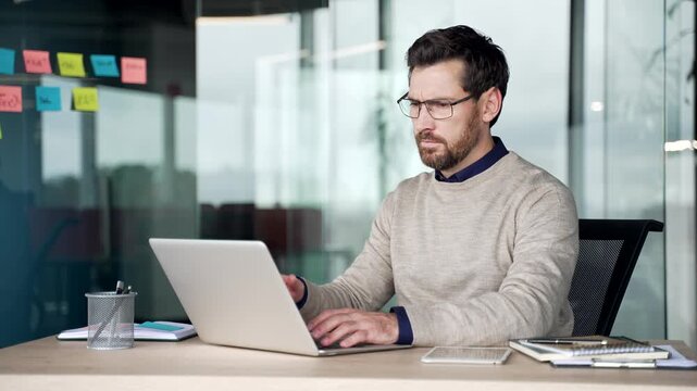 Frustrated businessman struggling with laptop while sitting at desk in office. Angry mature man reacts with irritation after problem on computer, feeling stressed and annoyed during work at workplace.