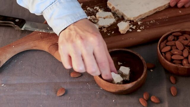 A person's hands carefully put pieces of hard white almond turron into a wooden bowl next to bowl of whole almonds, cutting board and a kitchen knife on a grey tablecloth