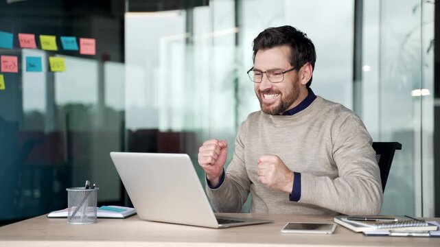 Excited businessman watching online auction on laptop at desk in modern office. Focused mature professional reacts with joy after winning bid, celebrating success and showing enthusiasm at workplace.