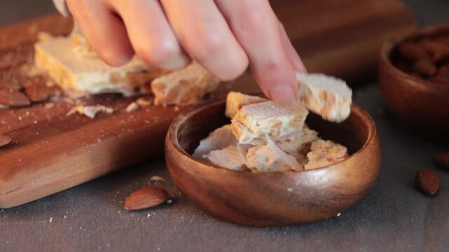 A person's hands carefully put pieces of hard white almond turron into a wooden bowl next to bowl of whole almonds, cutting board and a kitchen knife on a grey tablecloth