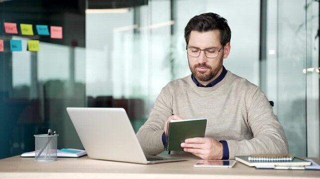 Businessman watching video conference on laptop while taking notes in notebook at desk in modern office. Focused mature professional listening to remote online training during work at workplace.