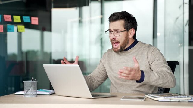 Angry businessman arguing during video call on laptop while sitting at desk in office. Frustrated mature professional having heated online discussion, reacting emotionally during remote conversation.