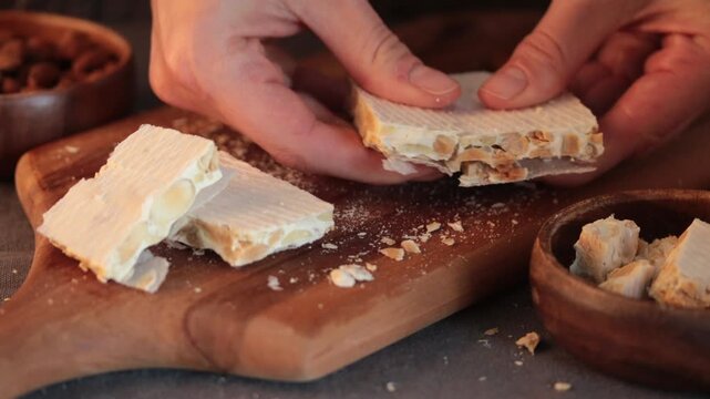 A close-up video of the moment a person's hands break a piece of traditional hard Spanish turron, filled with whole almonds, on a rustic wooden cutting board during a festive season