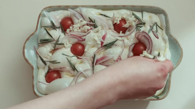 Hands adding shredded cheese to raw focaccia dough in ceramic baking dish already topped with cherry tomatoes, red onion, and rosemary