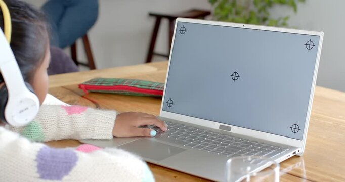 School-age girl with headphones clicking laptop at home desk for calibration, copy space