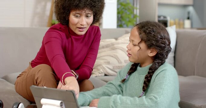 African American mom pointing at tablet at home guiding daughter tapping screen writing homework