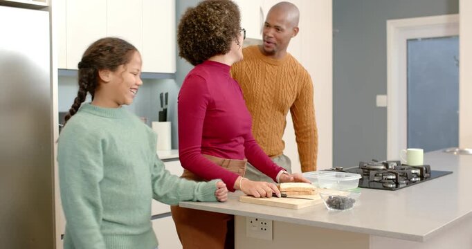 Diverse family girl entering kitchen hugging mom father grabbing mug slicing bread adding berries