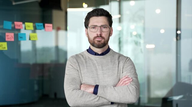 Serious confident businessman standing with crossed arms in modern glass office. Mature professional wearing glasses and sweater looking at camera with focused expression and leadership authority.
