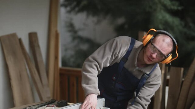 A focused carpenter, wearing protective ear muffs and safety glasses, expertly cuts a piece of wood on a table saw.