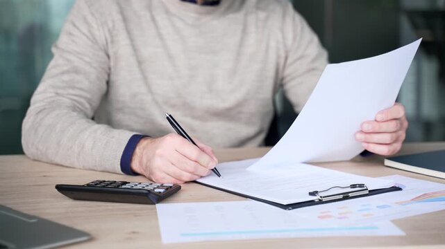 Close up of businessman hands filling document with pen while holding paper at desk in modern office. Mature professional writing notes, completing paperwork and focusing on details during work.