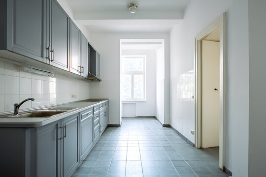 An empty kitchen featuring grey cabinets, a sink, and tiled walls/floors. A window and open doorway provide natural light