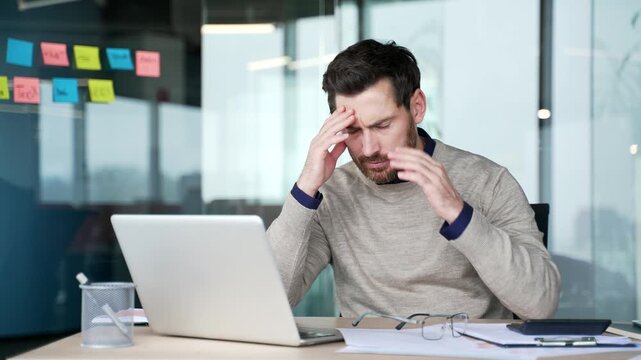 Exhausted businessman working on laptop and paperwork at desk in modern office. Frustrated mature professional facing difficulties, feeling despair and disappointment during stressful work process.