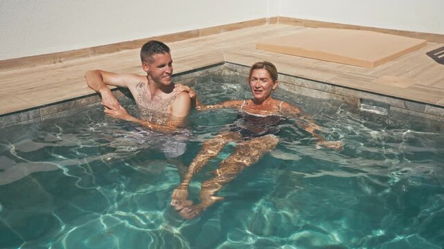 High angle view of smiling couple relaxing and talking in clear water of private swimming pool. Younger man and mature woman enjoying summer vacation, leisure, lifestyle and connection concept.