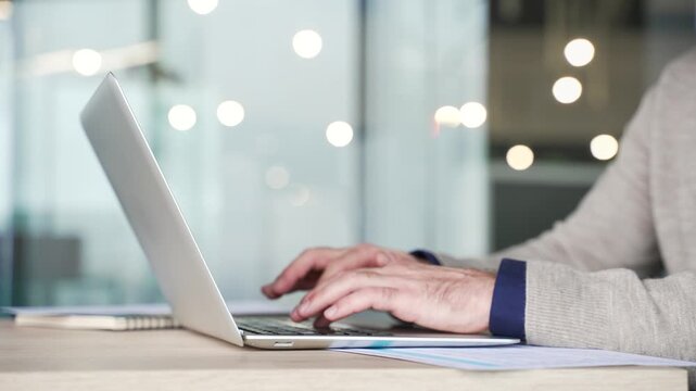 Close up of businessman hands typing on laptop keyboard at desk in modern office. Mature professional working on computer, entering text, managing information during daily business tasks at workplace.