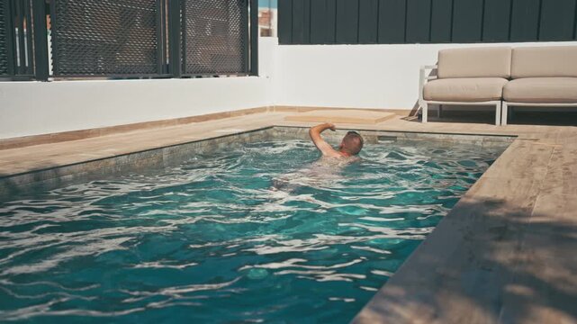 Young man swimming backstroke in private swimming pool at luxurious villa, relax by edge while enjoy summer vacation in beautiful and exclusive hotel resort