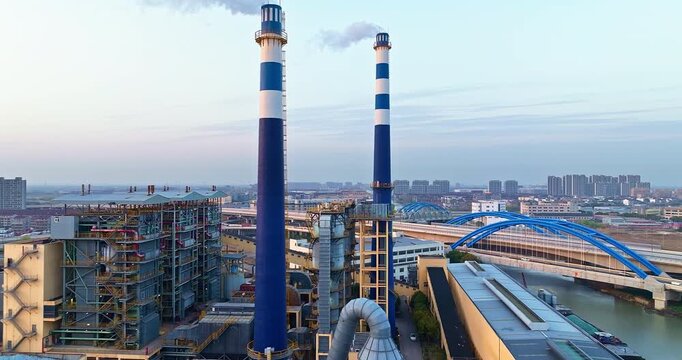 Industrial plant with tall smoking chimneys and modern architecture near city river under clear morning sky.