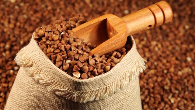 Close up of a small burlap bag with buckwheat groats and a wooden scoop on a scattered buckwheat background.