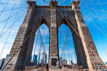 Obraz premium Symmetrical view of the Brooklyn Bridge pedestrian walkway leading toward the stone tower, framed by suspension cables and city buildings under a clear blue sky in New York City, USA. 