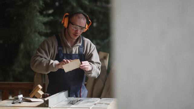 A focused carpenter wearing safety glasses and earmuffs precisely cuts wood on a table saw. He is diligently working on a woodworking project.