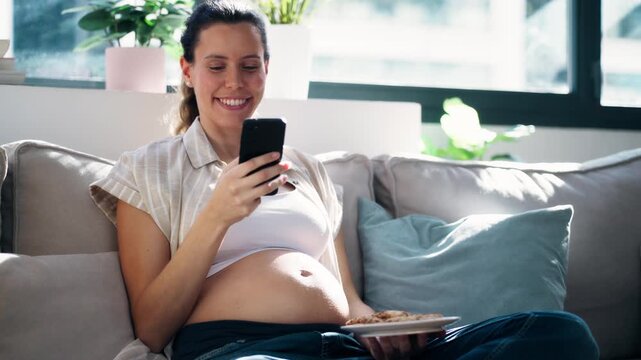 Video of pregnant woman using smartphone while eating a cookies sitting on the couch at home