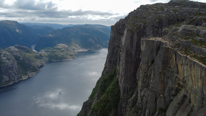 Obraz premium A fjord in Norway is surrounded by steep green slopes, with tourists walking along a winding path high above the deep blue water. Travel brochures for Scandinavian destinations, adventure tourism