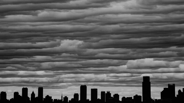 Dramatic city skyline under layered clouds, black silhouette of buildings stretches across low horizon, heavy textured cloud bands form ominous monochrome backdrop, looming storm suggests tension