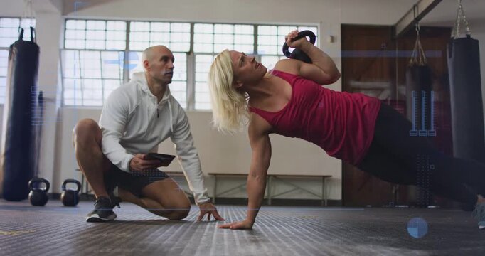 Vertical video: Client reaching kettlebell and lifting overhead in gym while trainer timing form
