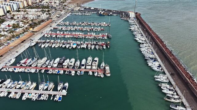 Boats are docked at the harbor. The water is calm. In the background, there are buildings and a breakwater. The scene shows a busy marina filled with different vessels.