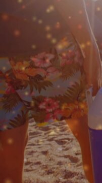 Pulling camera showing man in floral shorts, gray tank top walking on sand, holding blue cooler