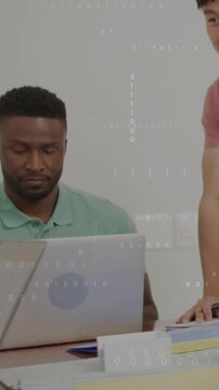 Vertical video: Seated man typing at laptop while standing man leaning in showing tech data overlay