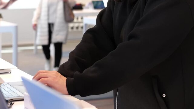 A close-up shot of a male customer in a black pullover trying out a laptop keyboard. The setting is a bright, white retail showroom for technology. He is carefully checking the keys and the device's f