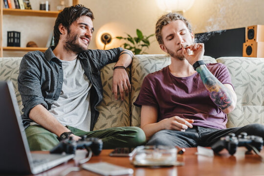 Two guys relaxing at home, smoking cannabis cigarettes, sitting on sofa, enjoying indoor leisure, personal hobby, lifestyle activity, casual home environment and modern lifestyle, addiction concept