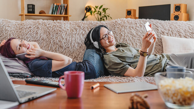 Two girlfriends lying on the sofa at home, enjoying music with headphones and scrolling through their mobile phones, embracing relaxation, digital connection, and cozy moments together.