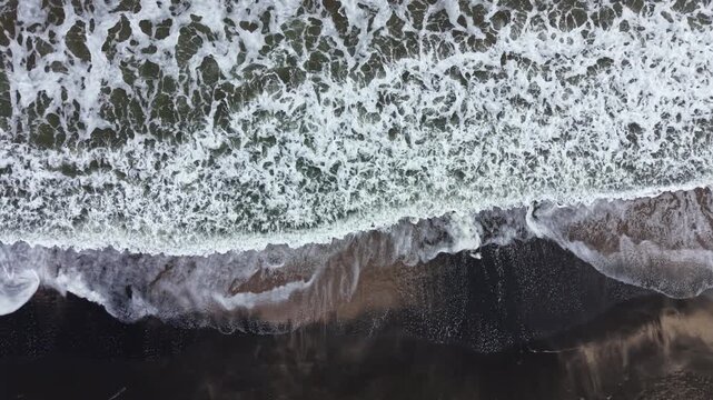 Waves break along the shore, forming white foam as they meet the sand. The tide is rolling in, creating patterns on the wet ground. The scene shows movement and change in nature.