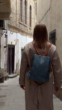 Back view of a female traveler walking through a narrow street in the old city of Baku, Azerbaijan, surrounded by aged stone walls and historic architecture, suitable for travel