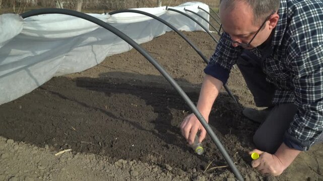 A gardener in a plaid shirt kneels by a bed, carefully adding fertilizer to the soil, covered with protective fabric and curved plastic hoops for optimal plant growth conditions in a greenhouse