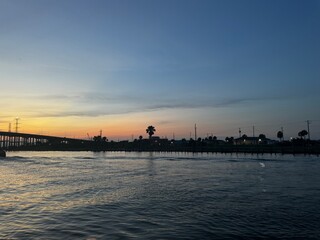 Fototapeta premium Calm water at sunset with a bridge, palm trees, and shoreline structures silhouetted against a soft gradient sky.