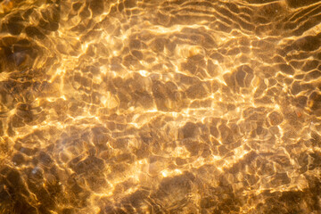 A clear stream of river water flows over a sandy-pebbly bottom on a sunny day. Sunlight creates golden wavy patterns on the surface and bottom of the reservoir. The water is shallow and has a warm gol © Alexei Vinnikov