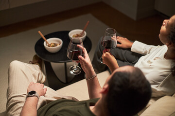 Young adult Caucasian man and young adult Black woman sitting together holding glasses of red wine, relaxing on sofa with bowls of food and chopsticks on small table in front