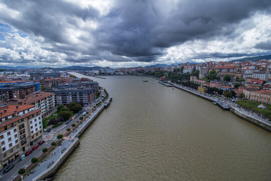 Gran plano general elevado que captura la desembocadura de la r&iacute;a hacia Bilbao, mostrando ambas m&aacute;rgenes, sus paseos mar&iacute;timos y edificios bajo un cielo dram&aacute;tico de nubes de tormenta.