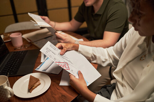 Young adult Black woman and young adult Caucasian man reviewing overdue bills at table, holding documents with past due notice, working together near laptop and sandwich