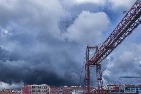 Composici&oacute;n que resalta la estructura met&aacute;lica del Puente Colgante cruzando el encuadre, con edificios residenciales de fondo y un cielo dram&aacute;tico cargado de nubes oscuras.