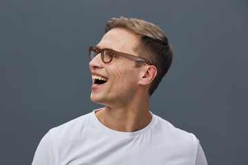 Obraz premium Joyful young man wearing glasses and a white t-shirt, smiling broadly with white teeth, looking to the side, against a plain gray background. Studio portrait, happiness, cheerful emotions.