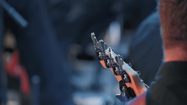 Musician playing bass guitar at a concert. Over the shoulder shot focuses on the headstock. Cool blue lighting and shallow depth of field create an atmospheric mood for a live music event